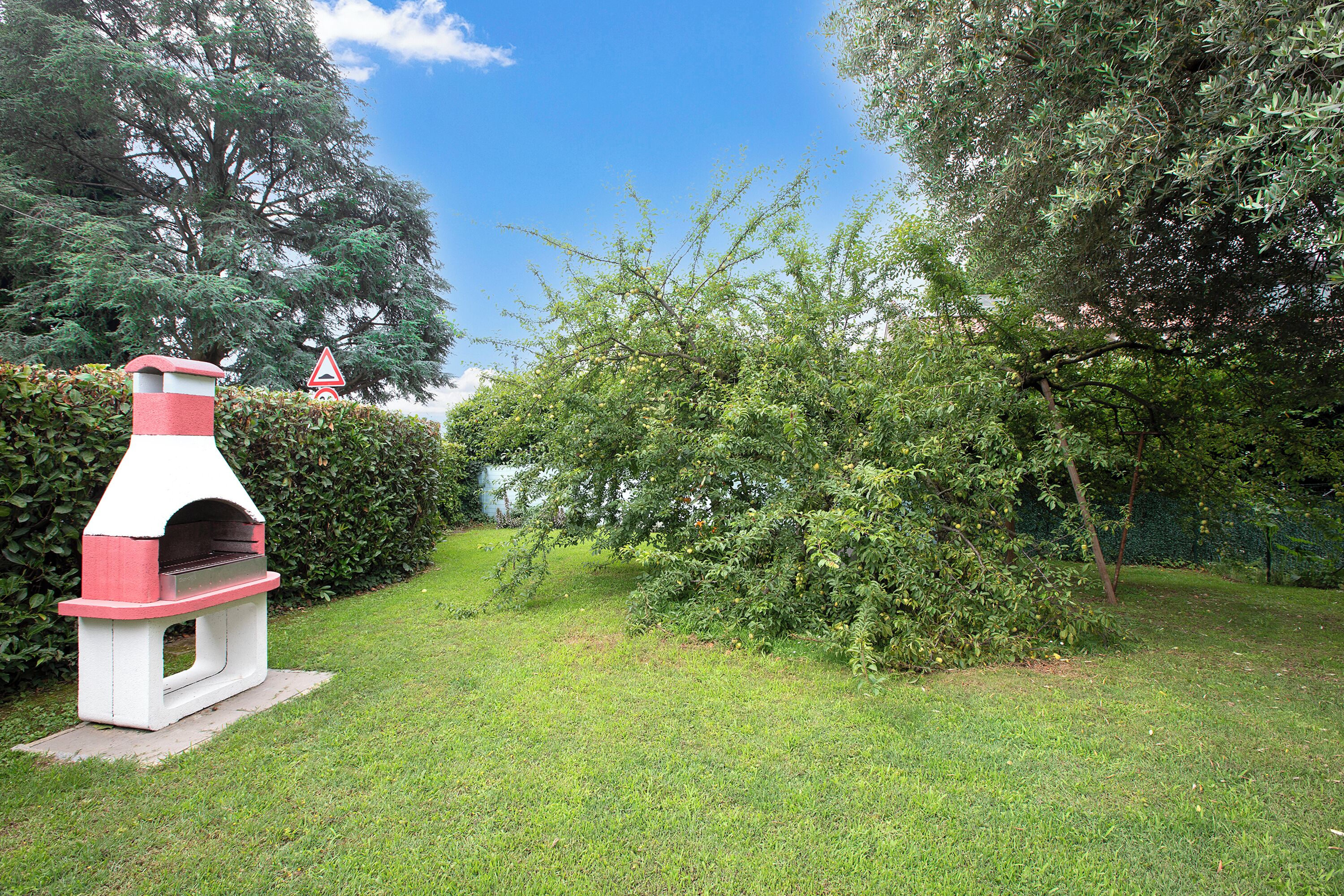 A red and white outdoor pizza oven in a grassy garden area with trees and a hedge in the background.