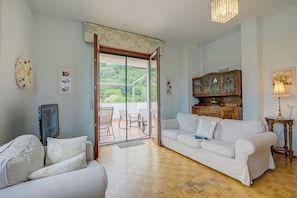 Ein Wohnzimmer mit einem weißen Sofa, einem Fernseher, einem Holzkabinett und einer Glastür, die auf eine Terrasse im Freien führt.