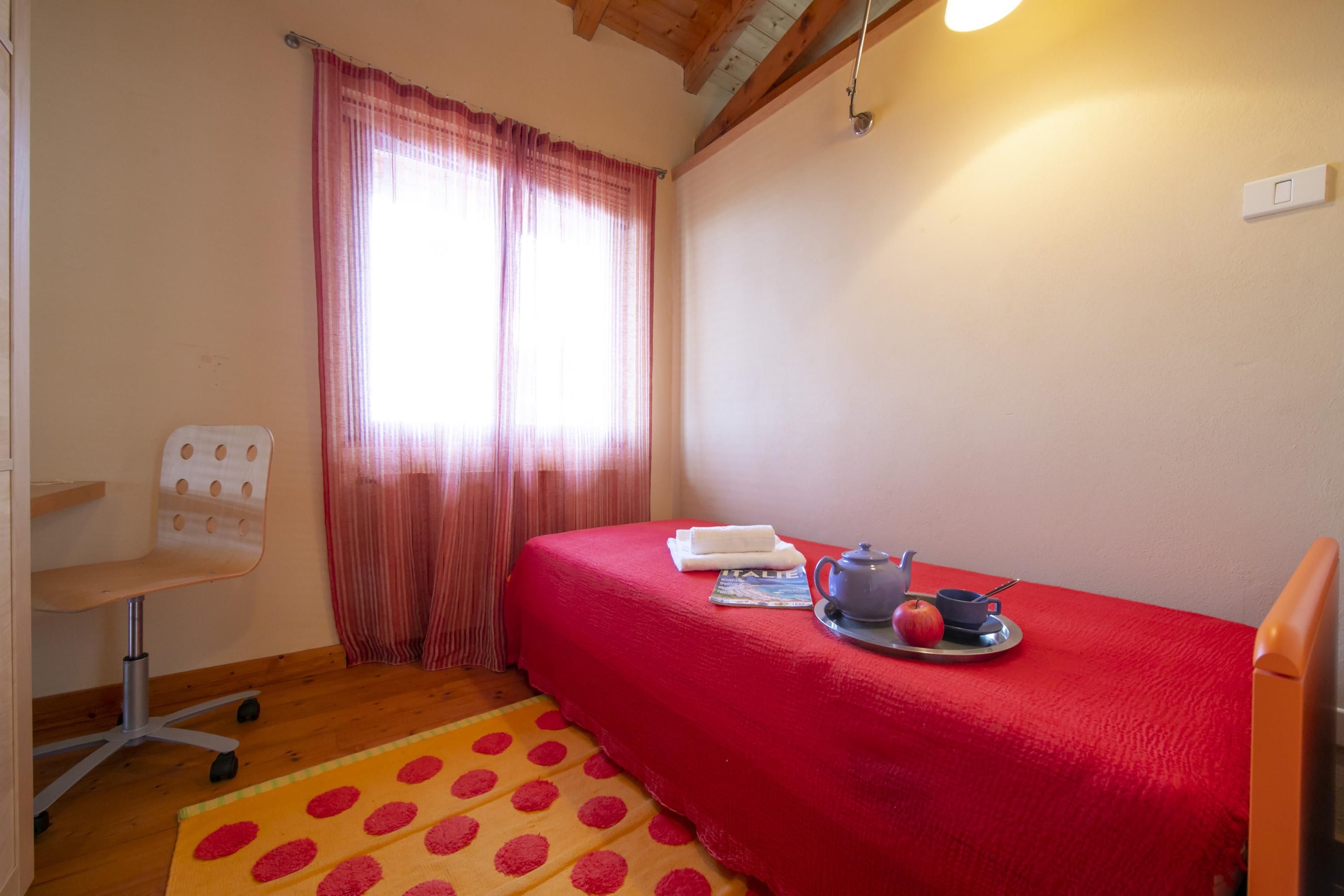 A bedroom with a bed, a chair, a window with curtains, a table with a tray, and a light fixture.