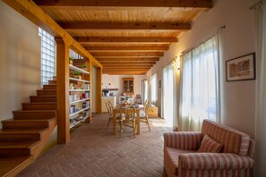 A spacious living area with a wooden staircase, a dining table with chairs, a sofa, and a bookshelf.