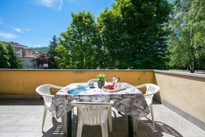 Een tafel met een tafelkleed met patroon, witte plastic stoelen en een kleine potplant op een balkon met een gele muur en bomen op de achtergrond.