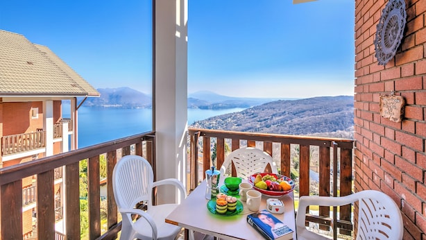 Een balkon met een ontbijttafel, inclusief een fruitschaal, een koffiepot en een boek. Het balkon biedt een prachtig uitzicht op een meer en bergen.