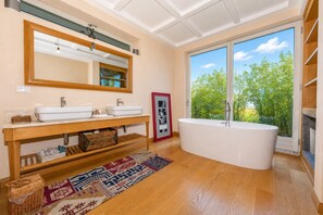 Casa de banho moderna com uma banheira independente, lavatório duplo, um espelho grande e vista para vegetação através de uma porta de correr de vidro.