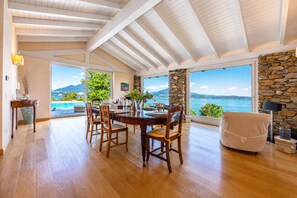 Un vaste espace dédié à la salle à manger, avec une table et des chaises en bois, de grandes fenêtres offrant une vue sur la mer et un mur en pierre.