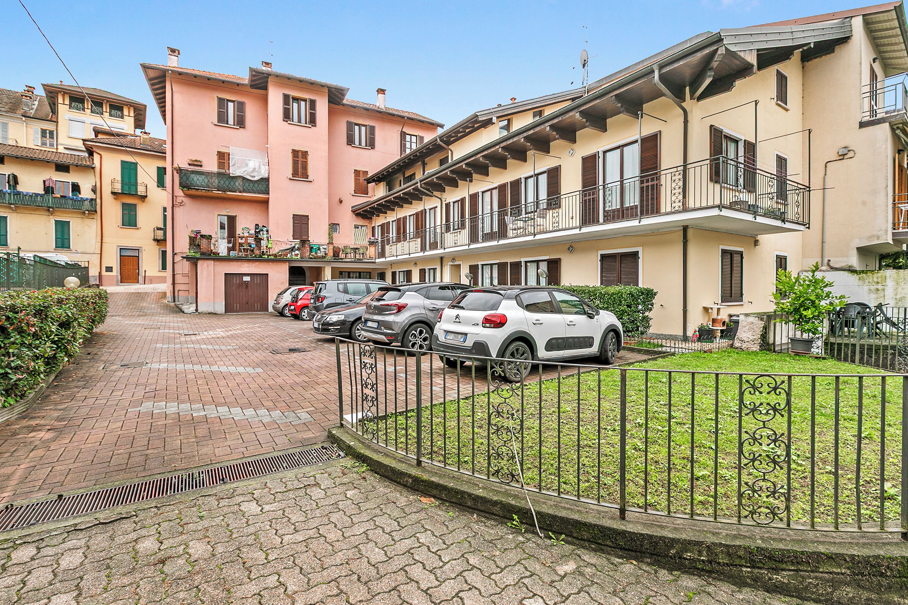 Une rue résidentielle bordée de voitures garées, avec un trottoir pavé et une zone herbeuse délimitée par une clôture en métal.