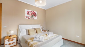 A neatly made bed with white linens and a beige and white checkered blanket, a bedside table with a lamp, and a floral painting on the wall. - Petalo Bianco 100m from lake (Baveno)