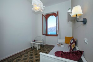 A room with a bed, a window with an open view, a chair, a small table, and a wall-mounted lamp.