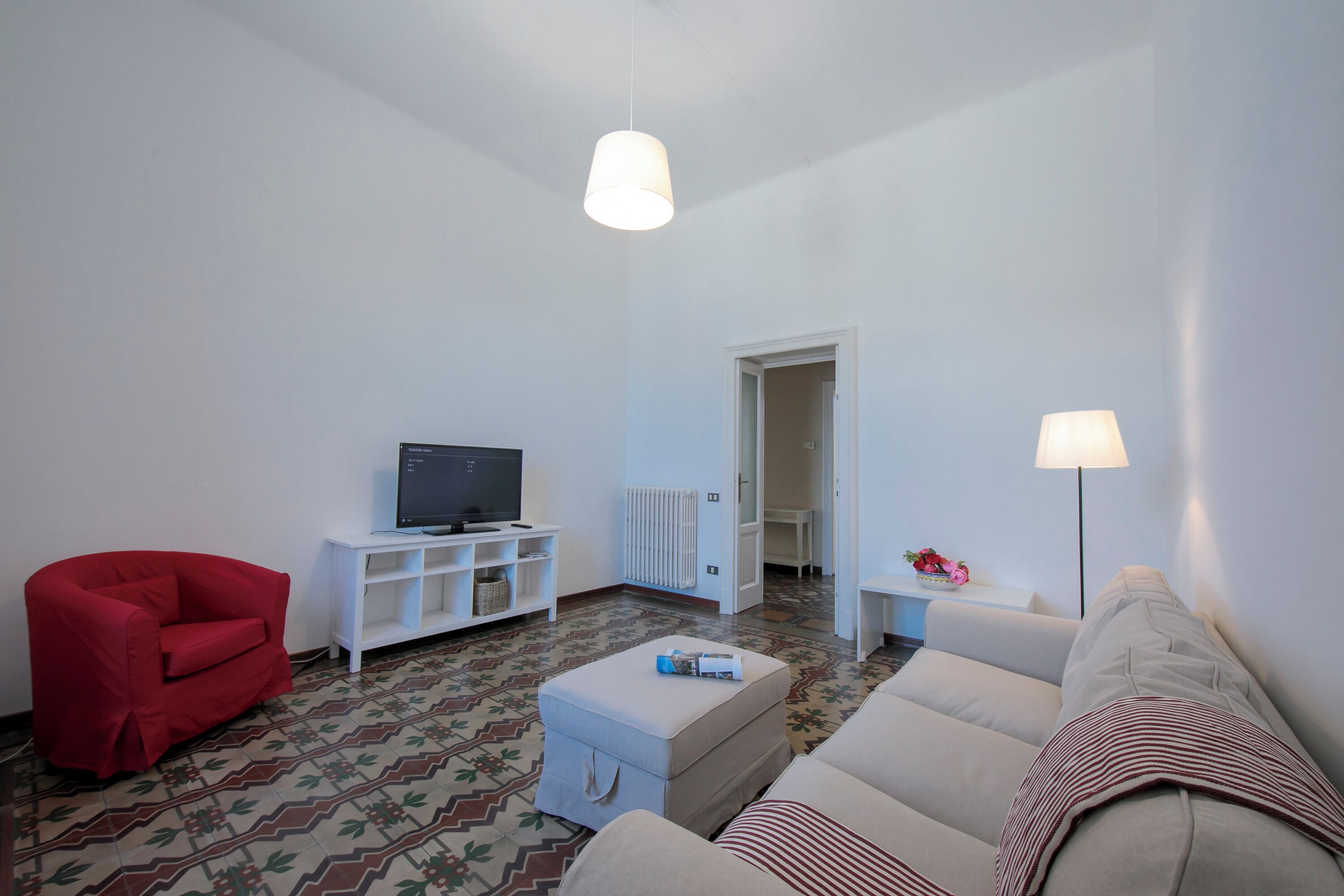 Un salon avec un fauteuil rouge, un canapé blanc, un pouf, une télévision, un meuble blanc et une lampe.