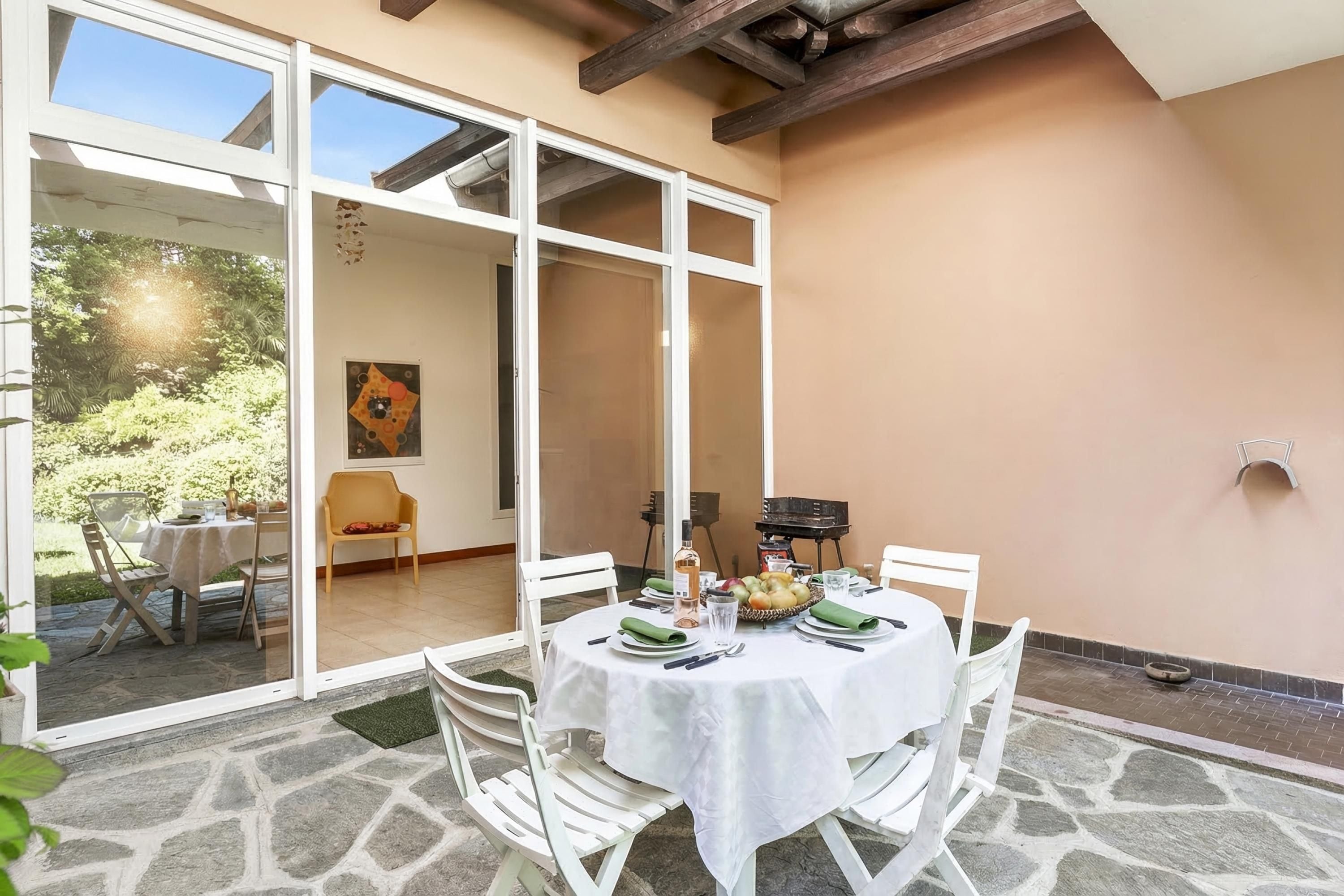 An outdoor dining area with a table set for two, surrounded by white chairs, a stone floor, and a glass door leading to a garden.