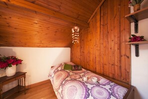 Una habitación acogedora con techo de madera, una cama con ropa de cama estampada, una mesita de noche con un florero y un estante con objetos decorativos.