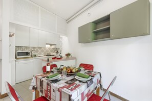 A compact kitchen with white cabinets, a microwave, and a dining table set with plates, glasses, and a fruit bowl. - Rododendro Rosso (Ispra)