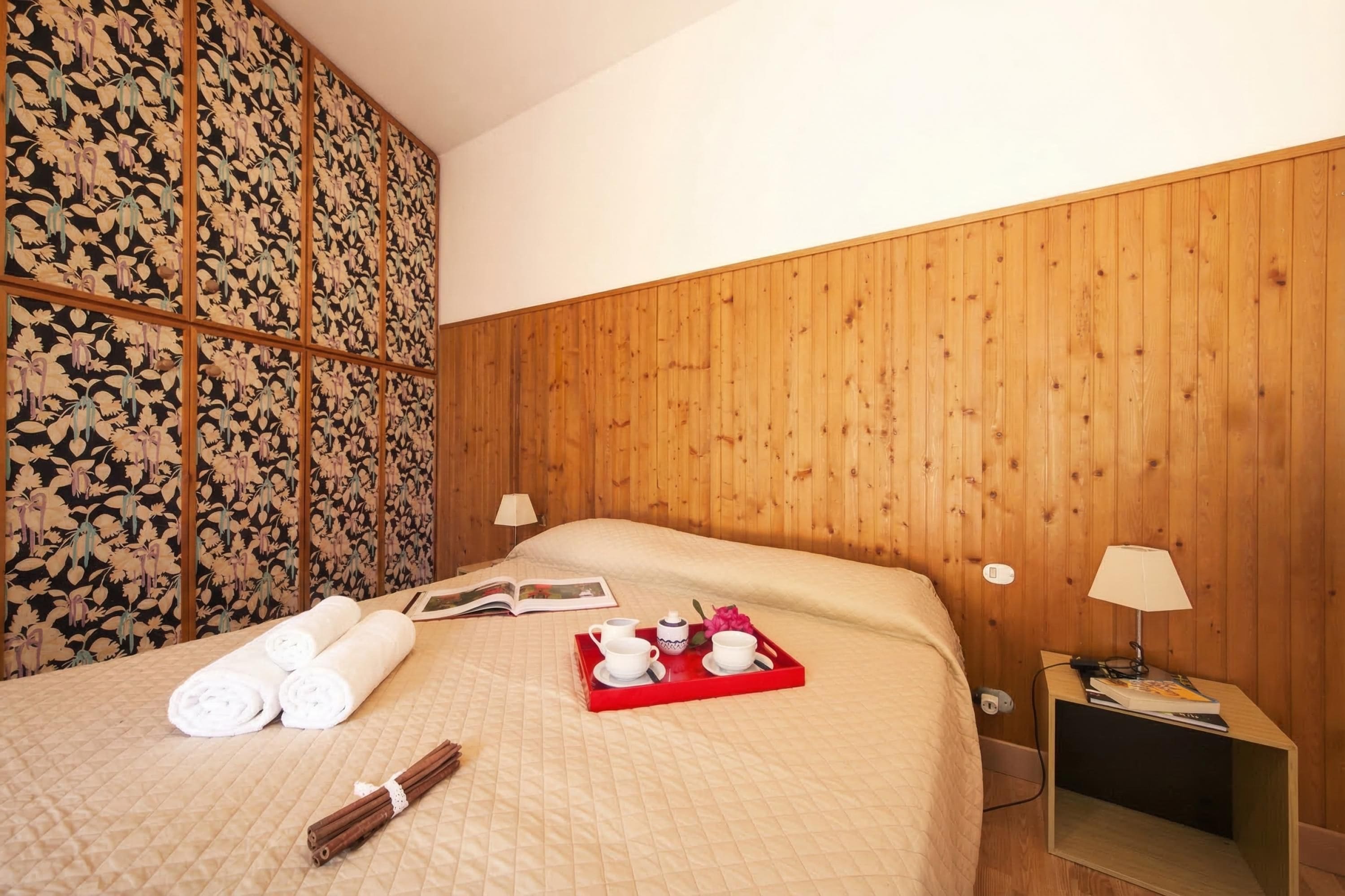A hotel room with a wooden headboard, a bed with a quilted bedspread, a bedside table with a lamp, and a tray with towels and a book.