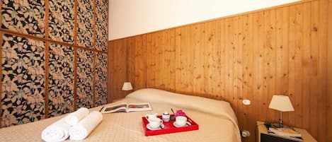 Een hotelkamer met een houten hoofdbord, een bed met een gewatteerde spreideken, een nachtkastje met een lamp en een dienblad met handdoeken en een boek.
