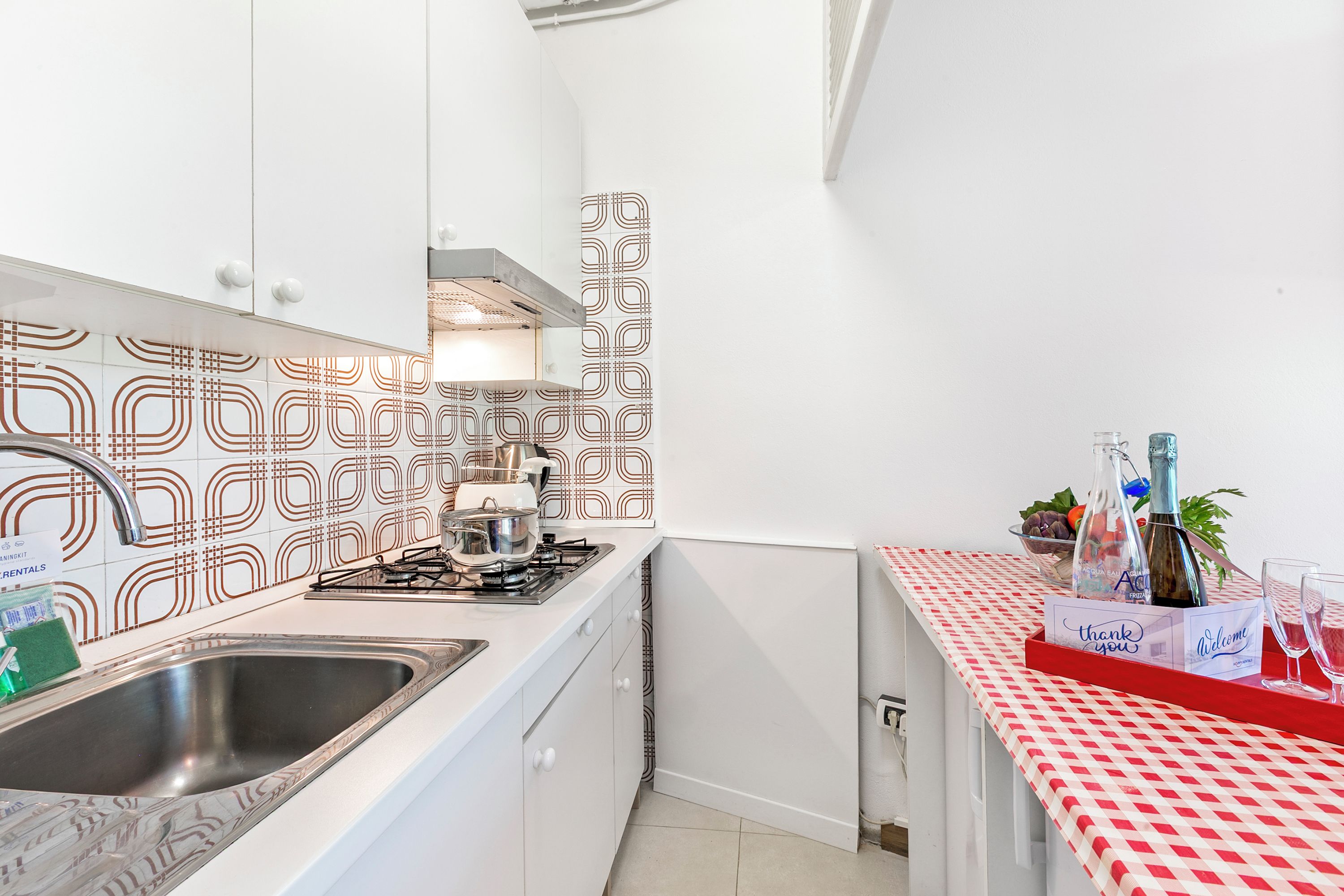 Et kompakt køkken med hvide skabe, en stålvask, en gasblus og mønstrede backsplash-fliser.