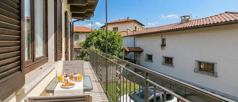 A balcony with a table set for breakfast, a car parked in the background, and a building with a tiled roof.