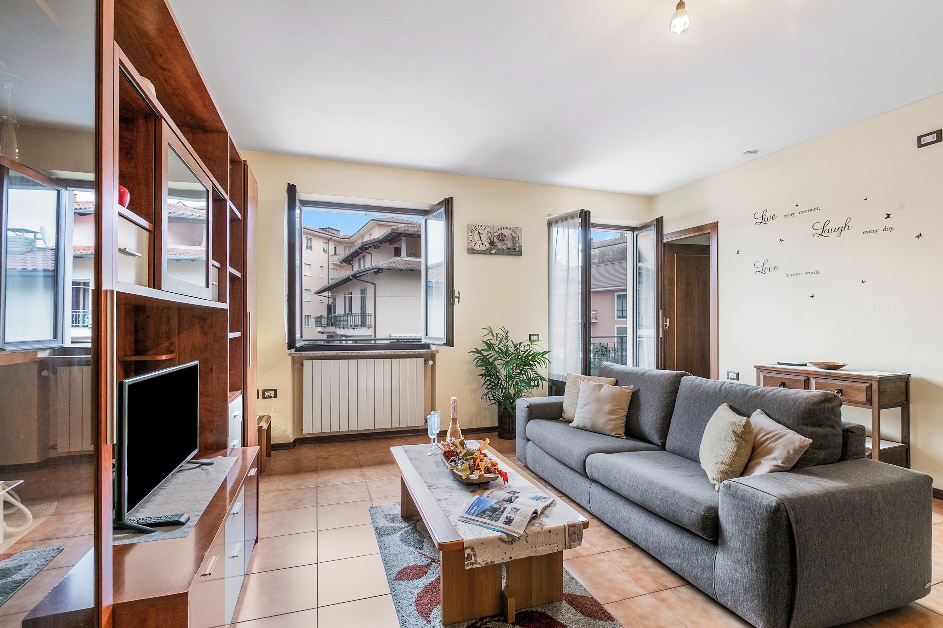 A modern living room with a grey sofa, a flat-screen TV, a wooden entertainment unit, and a large window offering a view of buildings.