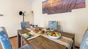 Ruokailutilassa on puinen pöytä, joka on katettu valmiiksi ateriaa varten. Pöydällä on kulho täynnä hedelmiä, juustoa ja leipätikkuja. Huoneessa on kaksi kukkapintaisella kankaalla verhoiltua tuolia, ja seinällä on taulu, jossa on kuvattu auringonlasku niityn yllä.