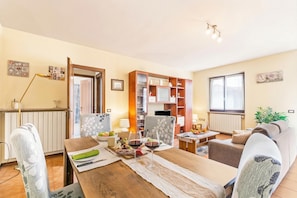 A cozy living room with a dining table set for two, a sofa, a bookshelf, and a window with curtains.
