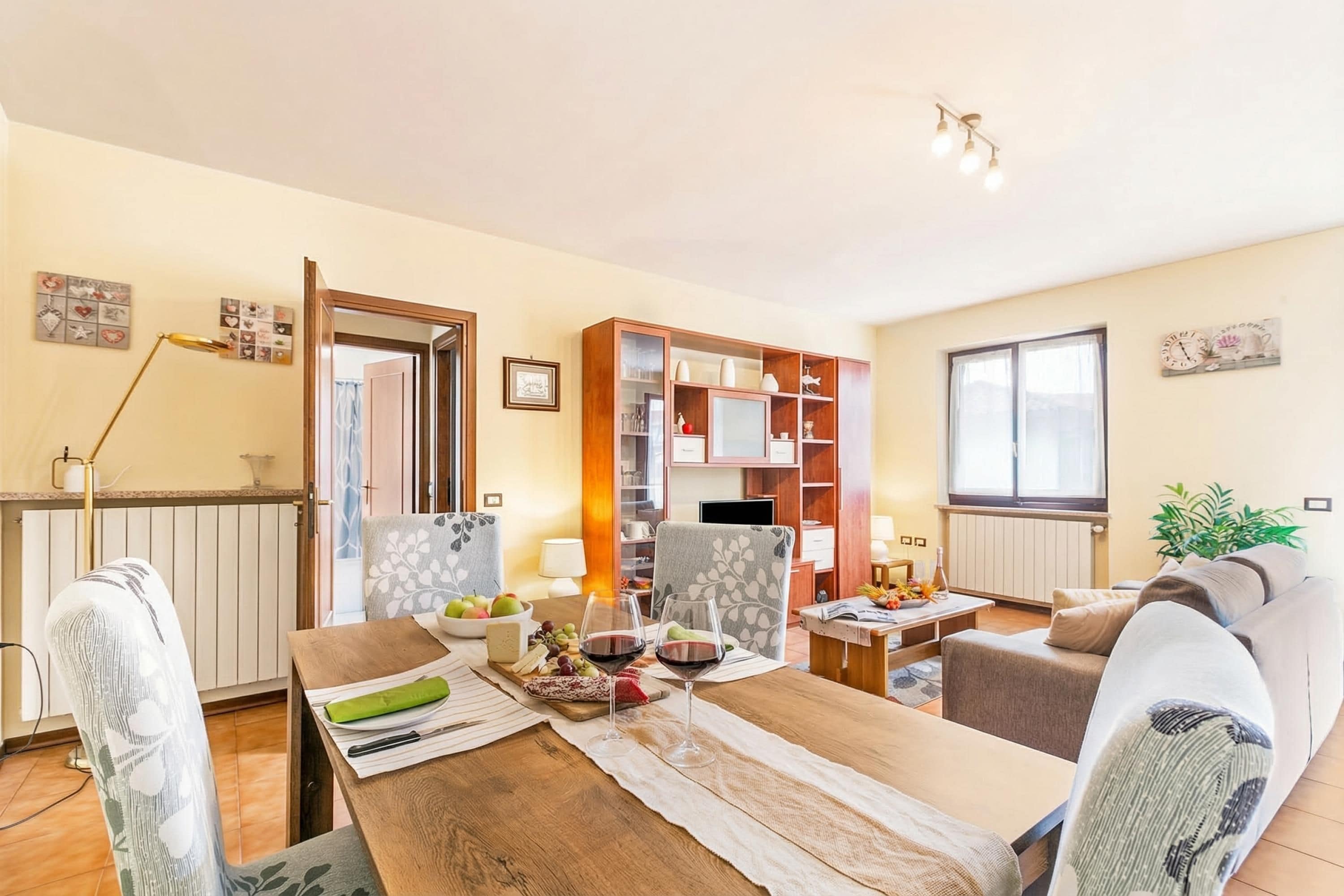 A cozy living room with a dining table set for two, a sofa, a bookshelf, and a window with curtains.