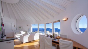 Een moderne, goed verlichte kamer met een houten vloer, witte meubels, grote ramen met uitzicht op bergen en een rond dakvenster.