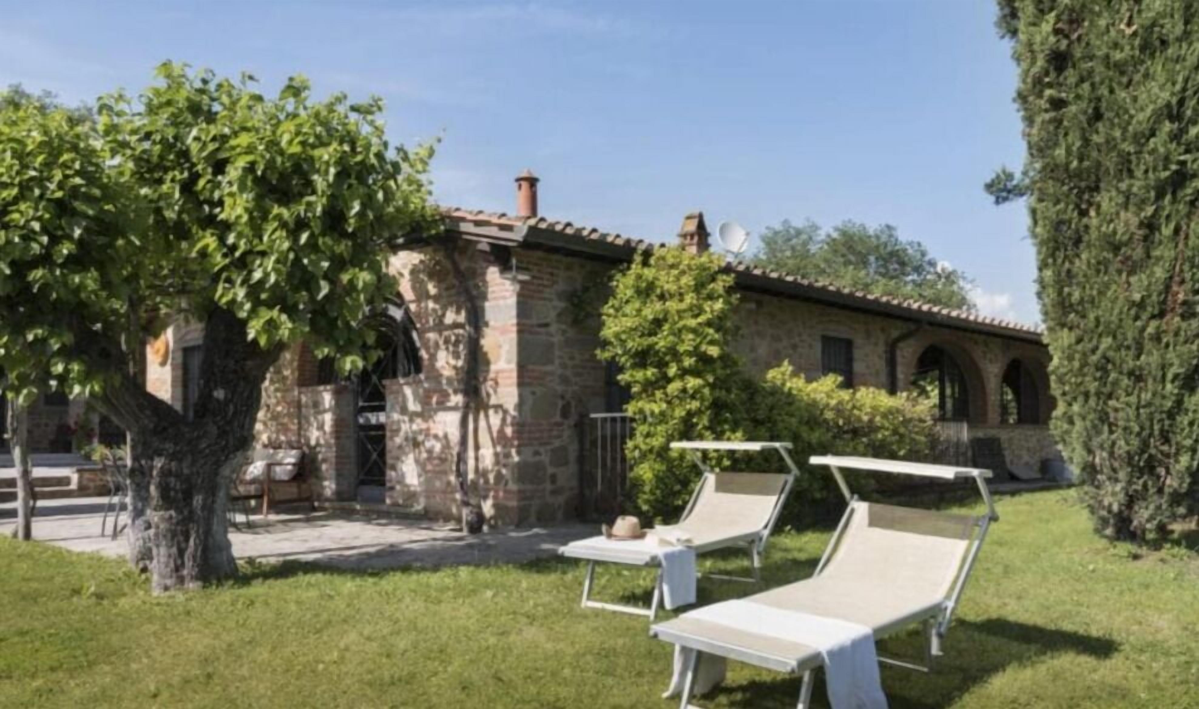 Un bâtiment en pierre avec un toit en tuiles, un arbre au premier plan et deux chaises longues sur une pelouse verdoyante.