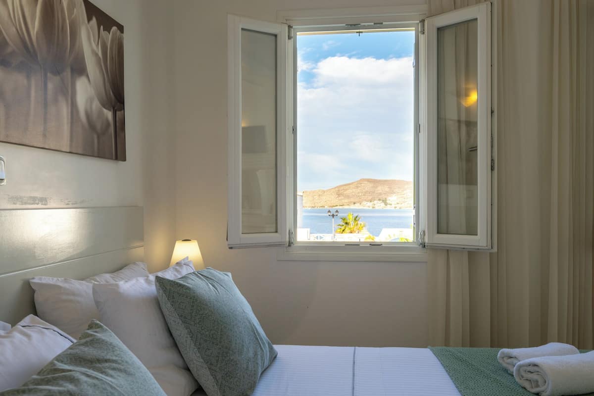 A hotel room with a bed, pillows, a lamp, and a window with a view of the sea and hills.