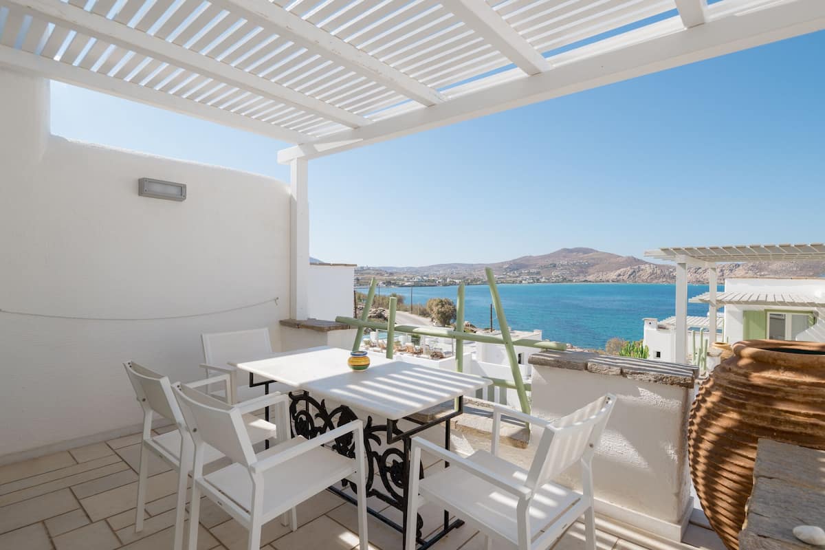 A balcony with a white table and chairs, overlooking a coastal view with mountains and the sea.