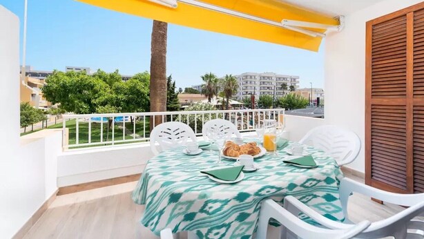 A balcony with a table set for breakfast, including croissants and orange juice, overlooking a residential area with palm trees and buildings. - Beautiful Apartment "Venecia" with Pool, Wi-Fi, Balcony & A/C (Alcudia)