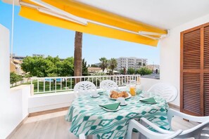 Un balcon avec une table dressée pour le petit-déjeuner, comprenant des croissants et du jus d’orange, donnant sur un quartier résidentiel avec des palmiers et des immeubles.