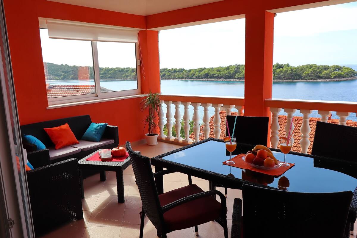 A balcony with a table and chairs, a sofa with cushions, and a view of a body of water and greenery.
