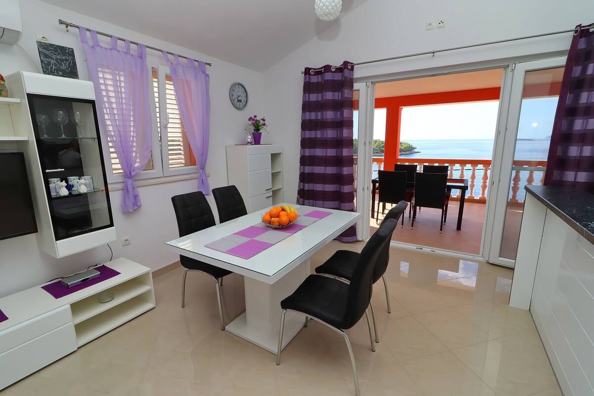 A modern dining area with a white table, black chairs, and a fruit bowl. There is a TV on a white cabinet, a white shelf with glassware, and a view of the sea through an open door.