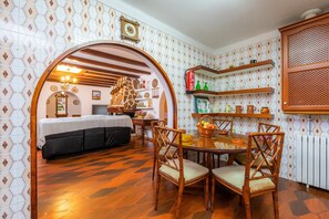 A room with a bed, a dining table with chairs, a kitchen area with shelves, and a patterned wall.