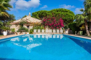 Une piscine avec un bâtiment en pierre en arrière-plan, entourée d’une végétation luxuriante et de plantes en fleurs.