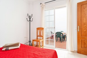 Un dormitorio con colcha roja, una silla de madera, perchero y puerta corrediza de vidrio que da a un espacio exterior con mesa y sillas.