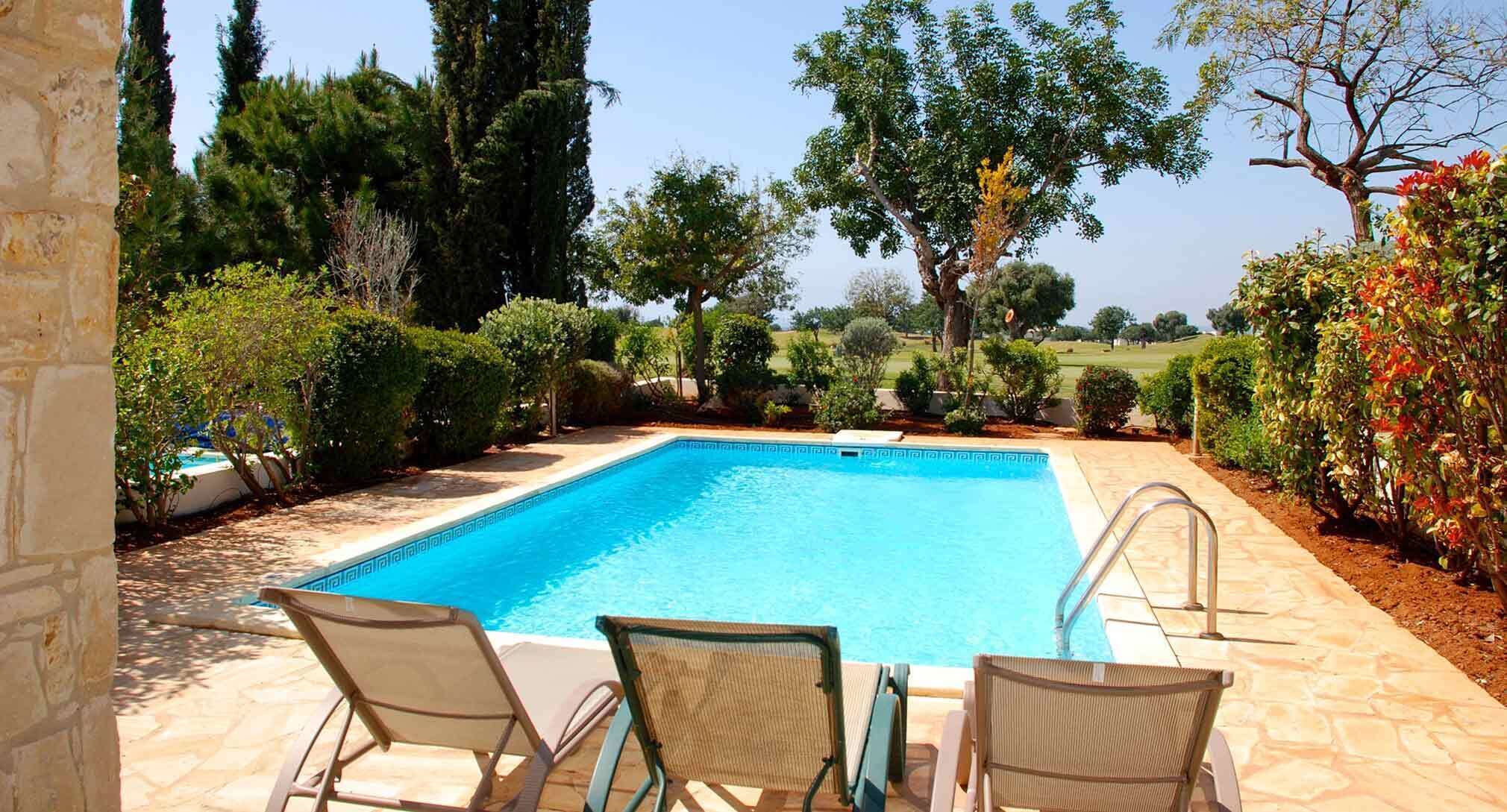 A rectangular swimming pool with a clear blue water surface, surrounded by a stone deck. There are two lounge chairs with woven backs and metal frames.