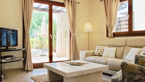 A living room with a television, a sofa, a coffee table, and a view of a garden through a glass door. - Villa HG30 - Destu (Kouklia)
