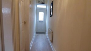 Un couloir étroit, avec une porte blanche au fond, un radiateur sur le mur de droite et une petite fenêtre au-dessus de la porte.