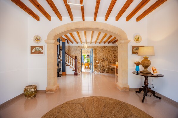 A hallway with a stone wall, wooden beams, a rug, a table with a lamp, and a vase.