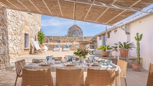 An outdoor dining area with wicker chairs and a table set for four, surrounded by potted plants and a stone wall. - Stone villa on the sea – Villa Can Cuarassa (Pollença)