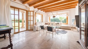 A spacious living area with a high ceiling, wooden beams, a sofa set, a dining table, and a large window offering a view of greenery.