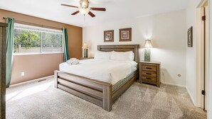 Une chambre à coucher comprenant un lit, une table de chevet, une lampe, un ventilateur de plafond et une fenêtre avec des rideaux.