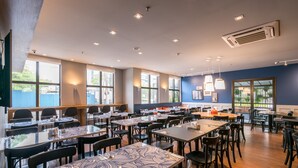Uma ampla sala de jantar com várias mesas e cadeiras, janelas grandes e luminárias no teto.