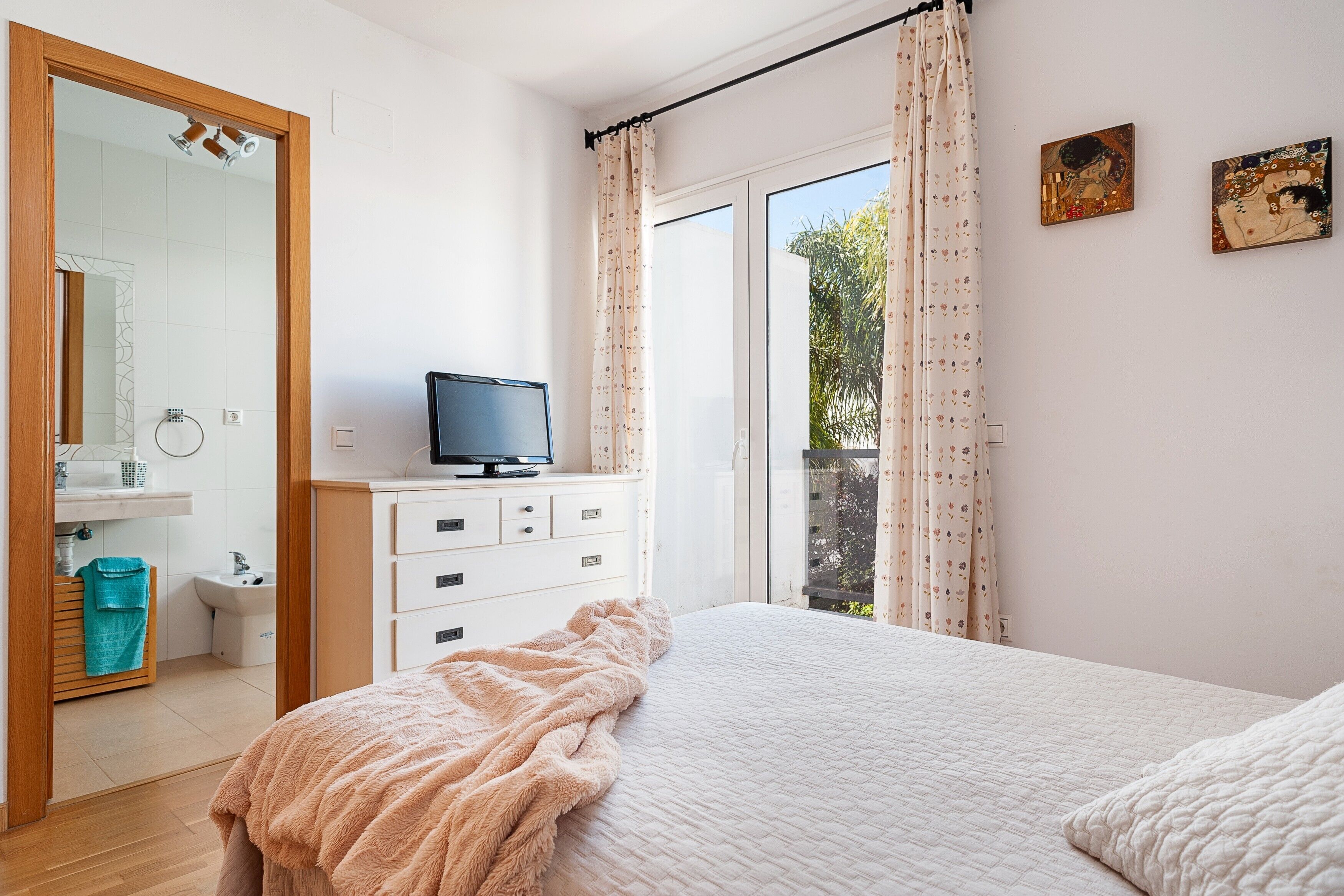A bedroom with a bed, a television on a dresser, a bathroom with a mirror, and a window with curtains.