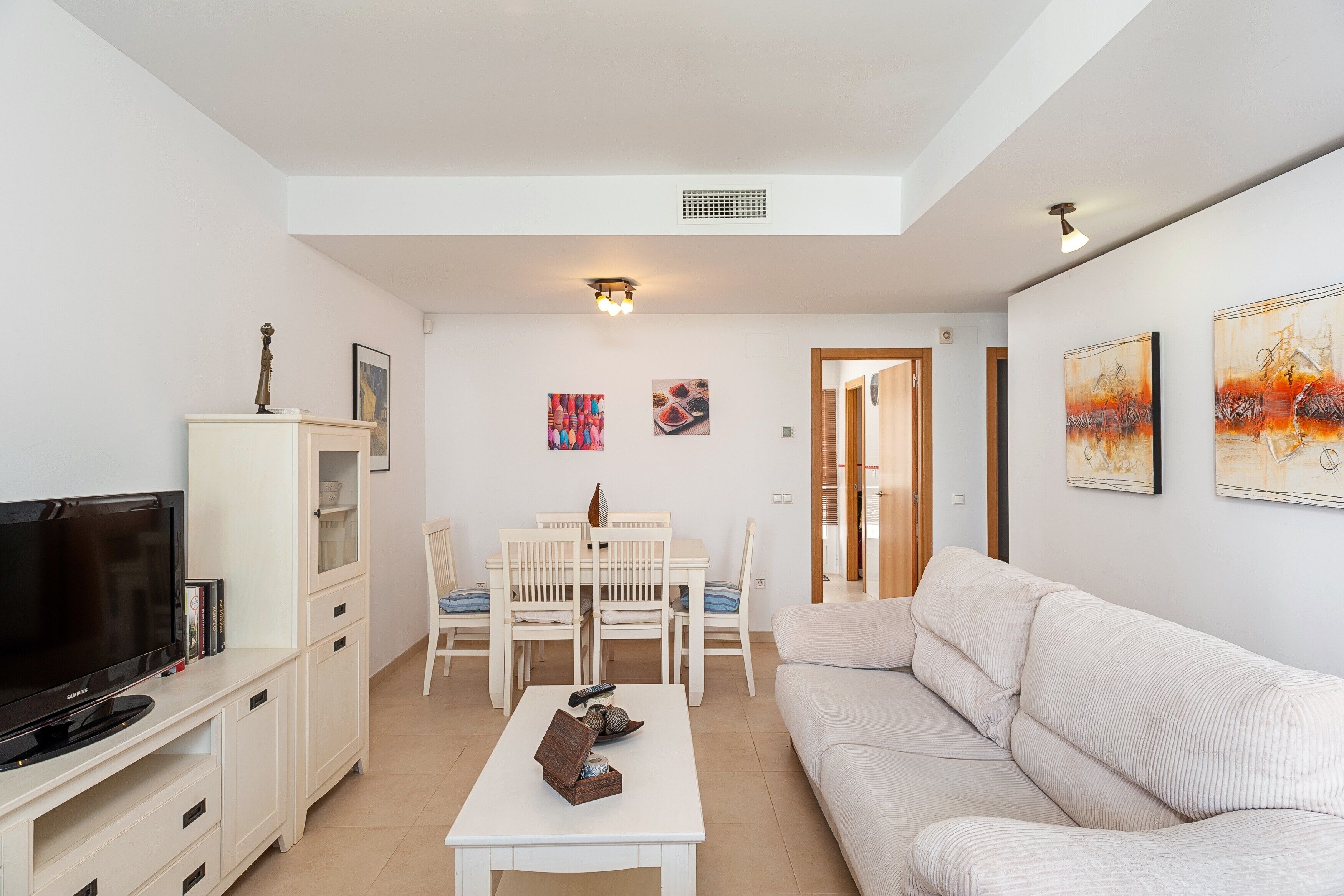 A living room with a TV, white furniture, a dining table, and artwork on the walls.