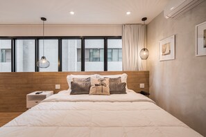 A modern hotel room with a large bed, a wooden headboard, a nightstand, a bedside table, a wall-mounted lamp, and a window with curtains.