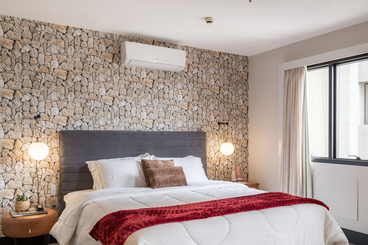 A modern bedroom with a stone wall, a large bed with white bedding and a red throw, two bedside lamps, and a checkered rug.