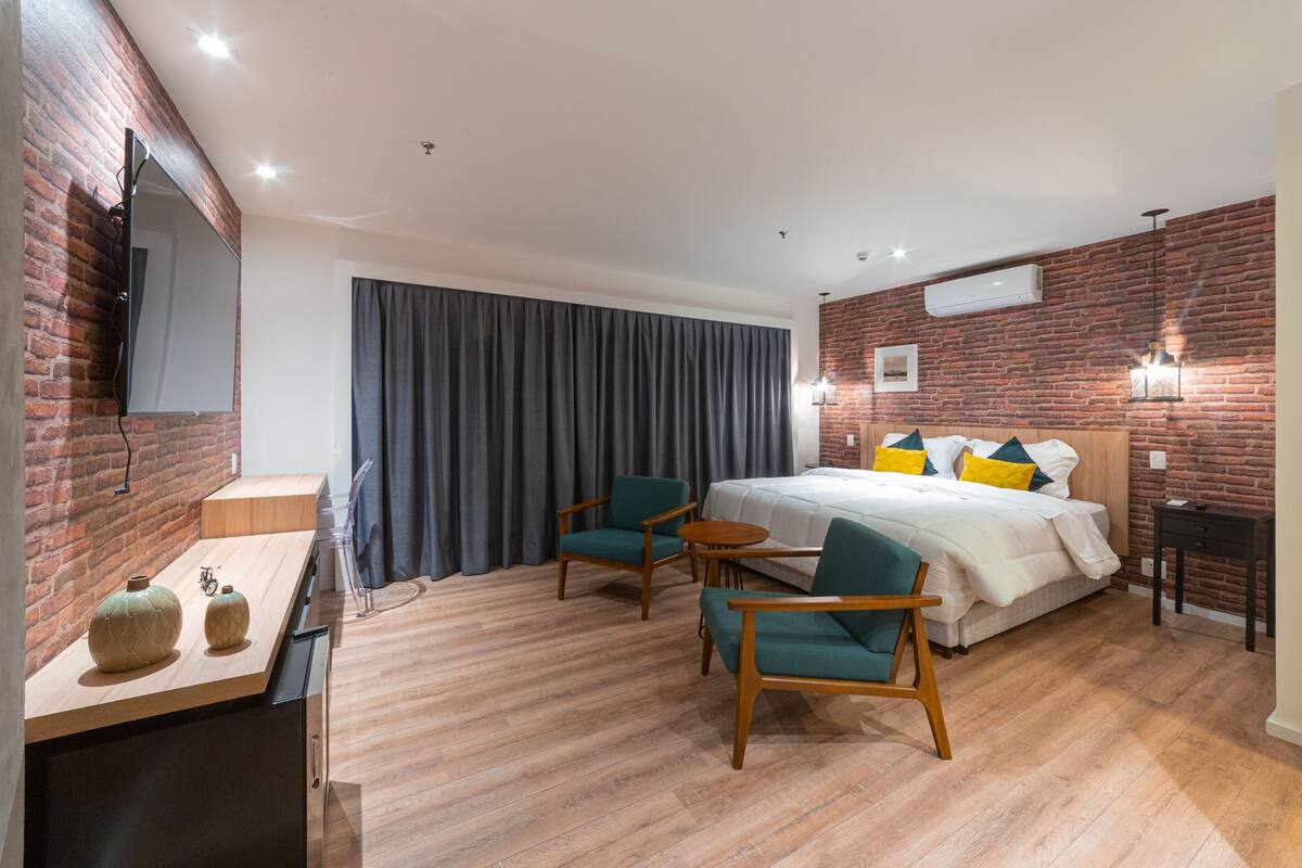 A modern hotel room with a brick accent wall, a large bed with colorful pillows, a desk with two vases, and a dining area with chairs and a small table.