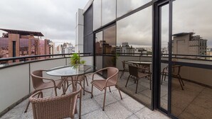A balcony with wicker chairs and a glass table, overlooking a cityscape with buildings and a cloudy sky. - Cyclinn Faria Lima Itaim (São Paulo)