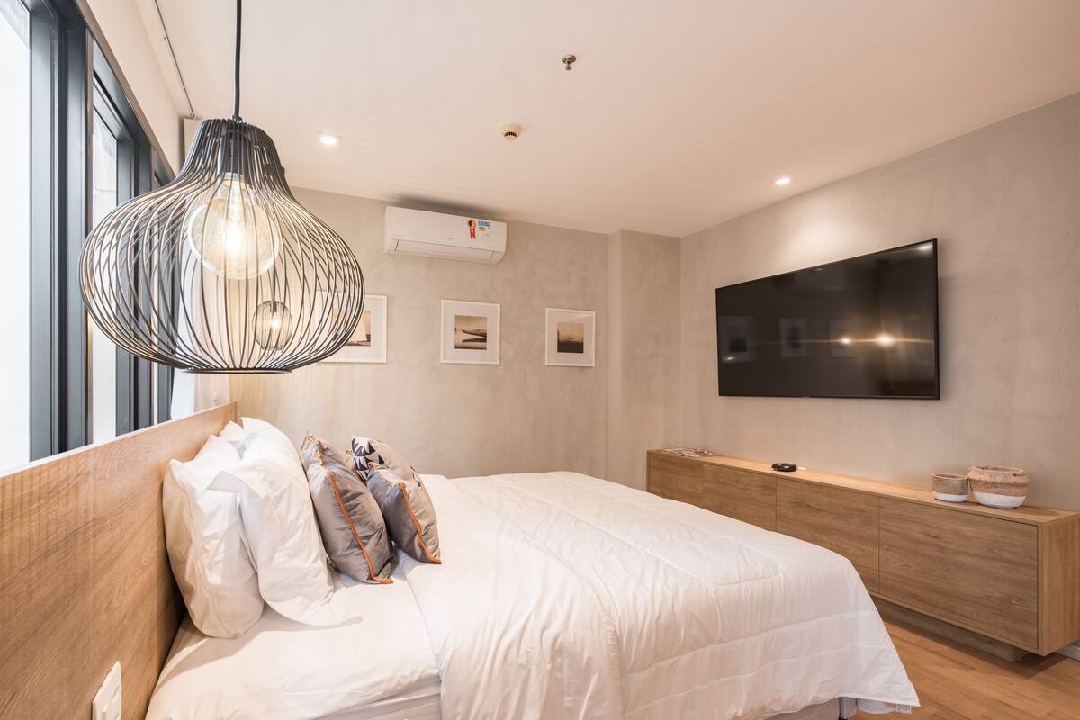A modern bedroom with a large bed, a hanging light fixture, a flat-screen TV, and a wooden nightstand.