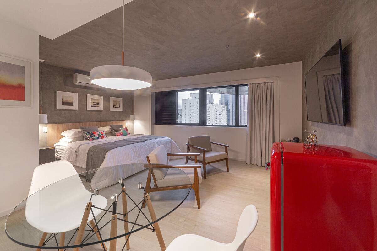 A modern bedroom with a bed, two chairs, a red cabinet, and a large window with a city view.